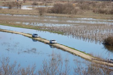 Hırvatistan 'ın Vrgorac kentinde 22,02,2026' lık manzara, sel sularının hakim olduğu sakin ama çarpıcı bir kırsal manzara sunuyor. Dar bir toprak yol olay yeri boyunca çaprazlama uzanır. Çevresindeki suyun üstünde kalacak kadar yükselir. İki araba gidiyor.