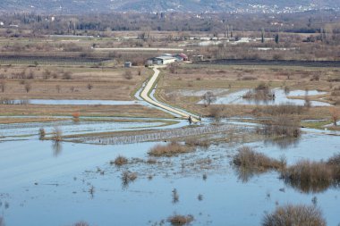 Hırvatistan 'ın Vrgorac kentinde 22,02,2026' lık manzara, sel sularının hakim olduğu sakin ama çarpıcı bir kırsal manzara sunuyor. Dar bir toprak yol olay yeri boyunca çaprazlama uzanır. Çevresindeki suyun üstünde kalacak kadar yükselir. İki araba gidiyor.