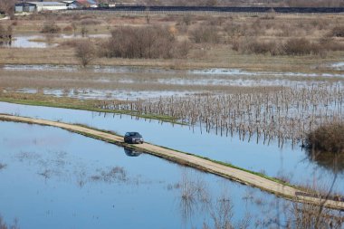Hırvatistan 'ın Vrgorac kentinde 22,02,2026' lık manzara, sel sularının hakim olduğu sakin ama çarpıcı bir kırsal manzara sunuyor. Dar bir toprak yol olay yeri boyunca çaprazlama uzanır. Çevresindeki suyun üstünde kalacak kadar yükselir. İki araba gidiyor.
