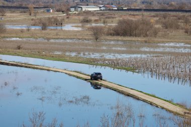 Hırvatistan 'ın Vrgorac kentinde 22,02,2026' lık manzara, sel sularının hakim olduğu sakin ama çarpıcı bir kırsal manzara sunuyor. Dar bir toprak yol olay yeri boyunca çaprazlama uzanır. Çevresindeki suyun üstünde kalacak kadar yükselir. İki araba gidiyor.