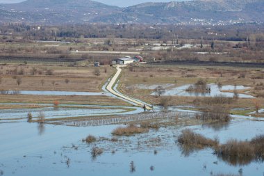 Hırvatistan 'ın Vrgorac kentinde 22,02,2026' lık manzara, sel sularının hakim olduğu sakin ama çarpıcı bir kırsal manzara sunuyor. Dar bir toprak yol olay yeri boyunca çaprazlama uzanır. Çevresindeki suyun üstünde kalacak kadar yükselir. İki araba gidiyor.