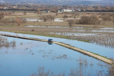 Hırvatistan 'ın Vrgorac kentinde 22,02,2026' lık manzara, sel sularının hakim olduğu sakin ama çarpıcı bir kırsal manzara sunuyor. Dar bir toprak yol olay yeri boyunca çaprazlama uzanır. Çevresindeki suyun üstünde kalacak kadar yükselir. İki araba gidiyor.