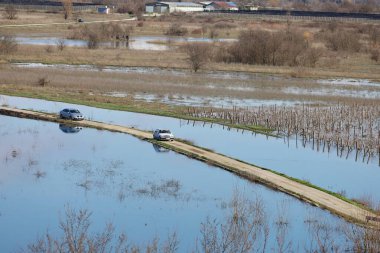 Hırvatistan 'ın Vrgorac kentinde 22,02,2026' lık manzara, sel sularının hakim olduğu sakin ama çarpıcı bir kırsal manzara sunuyor. Dar bir toprak yol olay yeri boyunca çaprazlama uzanır. Çevresindeki suyun üstünde kalacak kadar yükselir. İki araba gidiyor.