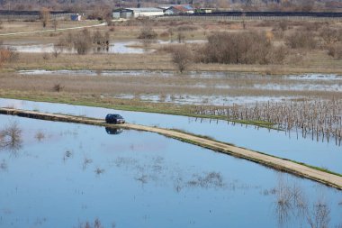 Hırvatistan 'ın Vrgorac kentinde 22,02,2026' lık manzara, sel sularının hakim olduğu sakin ama çarpıcı bir kırsal manzara sunuyor. Dar bir toprak yol olay yeri boyunca çaprazlama uzanır. Çevresindeki suyun üstünde kalacak kadar yükselir. İki araba gidiyor.