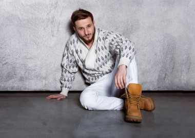 Beyaz kot pantolon ve hırka giyen, oturarak beton arka planda poz veren kendine güvenen genç bir adamın yakın portresi..