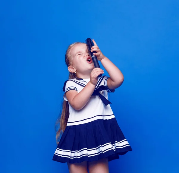 Schattig klein meisje met blauwe ogen, draagt een jurk, houdt een kam vast in plaats van een microfoon op een blauwe achtergrond. Plaats voor tekst. — Stockfoto