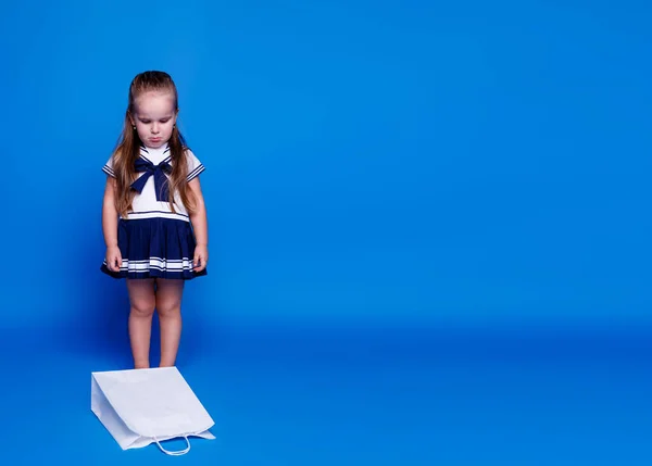 Ongelukkig klein mooi meisje, in een jurk, kijkend naar de witte boodschappentas zittend op de vloer, over blauwe achtergrond. — Stockfoto