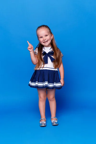 Schattig lachend meisje wijst met haar vinger omhoog, draagt een jurk, geïsoleerde blauwe achtergrond, studio opname. Verticaal overzicht. — Stockfoto