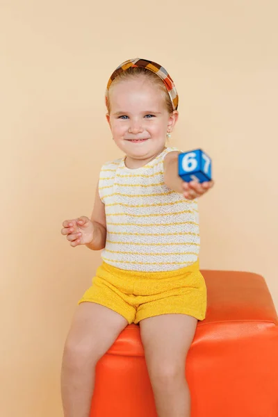 Menina muito sorridente, sentada na cadeira laranja, com números de blocos brinquedos, sobre fundo bege, olhando para a câmera. — Fotografia de Stock