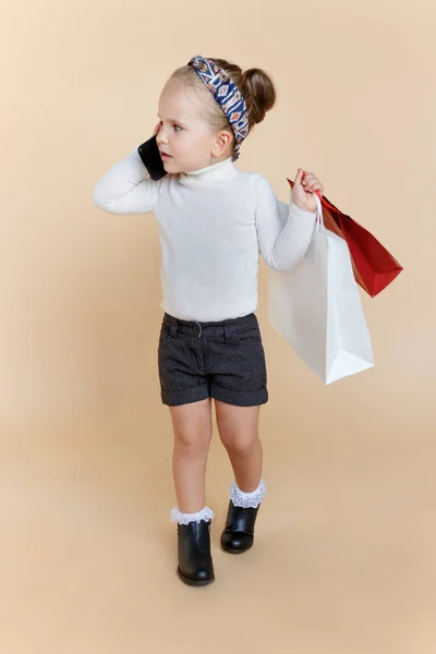 Schattig klein meisje in de herfst outfit met een shoppers tassen in de hand, praten op een mobiele telefoon, over beige achtergrond. — Stockfoto