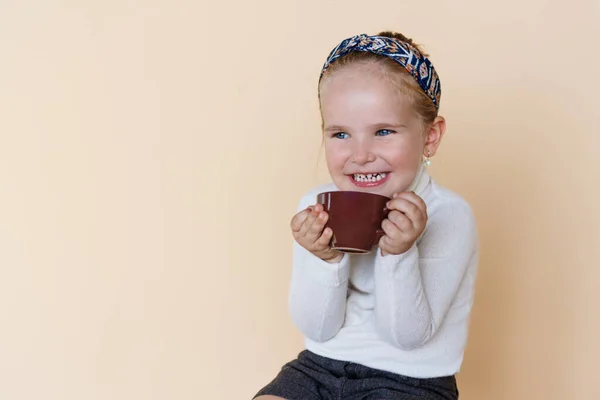 Portret van een klein schattig meisje zitten en glimlachen, met een kopje warme drank cacao genieten, over beige achtergrond. — Stockfoto