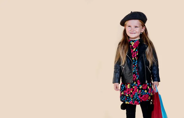 Modieus klein meisje met lang haar in leren jas en baret, met boodschappentassen, over beige achtergrond. — Stockfoto