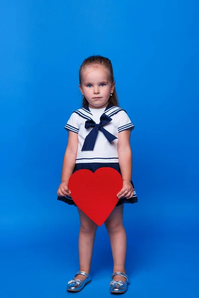 Doce, menina pensativamente criança 3 anos de idade vestindo vestido azul segurar na mão coração vermelho isolado no fundo da parede azul. — Fotografia de Stock