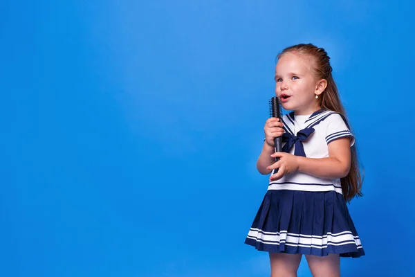 Söt liten flicka i klänning håller en kam i stället för en mikrofon på blå bakgrund. Plats för text. Horisontell vy. — Stockfoto