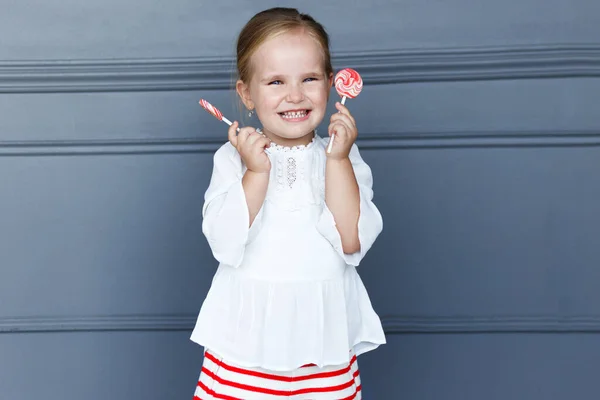 Mooi klein meisje met lolly, glimlachend en kijkend naar de camera, geïsoleerd op een grijze neoklassieke achtergrond. Kopieerruimte. — Stockfoto