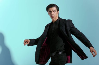 Genç adam stüdyoda tavır takınıyor, kameraya bakıyor, takım elbiseli, mavi arka planda soyutlanmış..