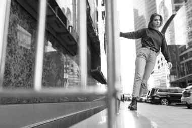 Siyah-beyaz, kısa sarı saçlı, mavi giysili, kot pantolonlu, çizmeli, kaba sokak duvarına karşı poz veren bir kız..