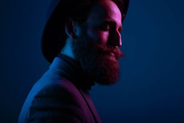 Şapka ve takım elbiseli sakallı bir adamın portresi. Stüdyoda profilden poz veriyor, mavi arka planda izole edilmiş..