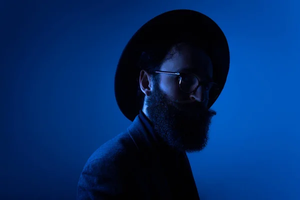 Vousatý muž v klobouku a obleku, pózování v profilu ve studiu, studiová světla, izolované na tmavomodrém pozadí. — Stock fotografie
