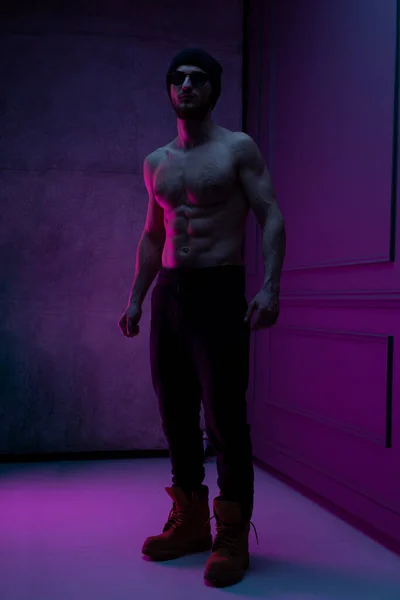 Full length portrait of a brutal man with bare torso posing in the studio with concrete wall with purple neon color. — Stock Photo, Image