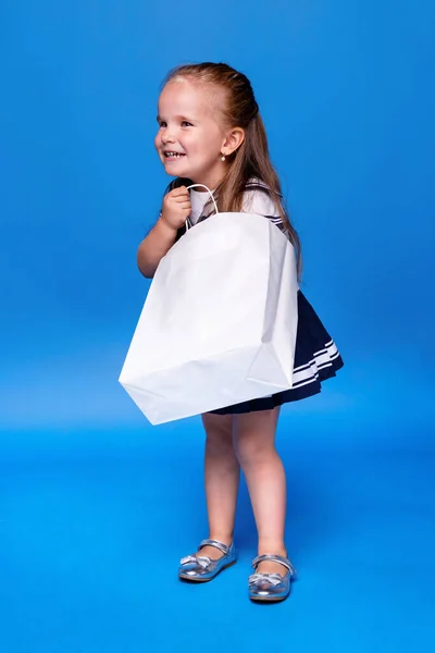 Glimlachend meisje met lang haar, in een jurk, kijkend in een witte boodschappentas, op een blauwe achtergrond. — Stockfoto