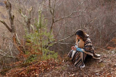 Kumral saçlı, pançolu genç bir kadın sonbahar ormanlarında yürür..