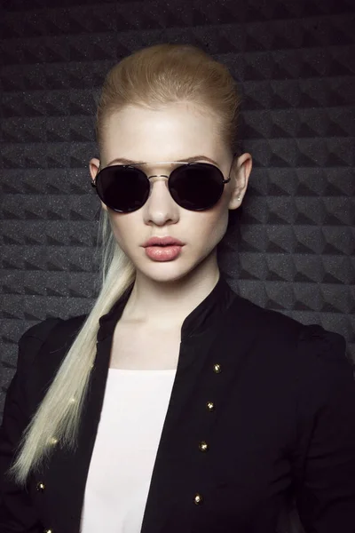 Szoros kép egy érzéki szőke modell smink és lófarok frizura, visel fekete kabát és napszemüveg. — Stock Fotó