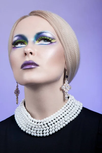 Portrait of a female model with blonde hair and a fashion make-up, looking up with pearl necklace and cross earrings.