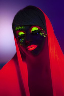 Kafasında turuncu eşarp ve renkli makyajlı genç bir kadın, gözleri kapalı, mor bir arka planı var..