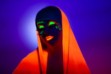 Artistik makyajlı, kafasında turuncu eşarp olan, gözleri kapalı, mor arka planda bir kadının ön görüntüsü..