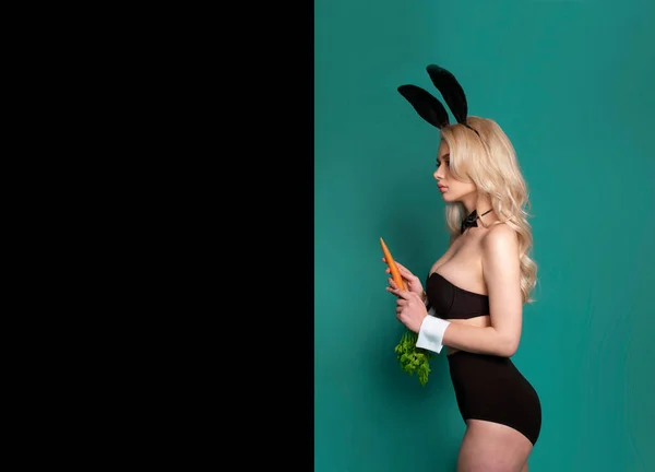 Profilporträtt av en sexig het blond-kvinnlig modell i svarta underkläder med kaninöron, med en morot i handen. — Stockfoto
