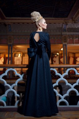 Uzun siyah bir elbise giymiş, arkası fiyonklu, zarif saç stili, taç ve küpe takan muhteşem sarışın bir manken..