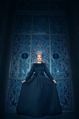 Uzun siyah bir elbise giymiş, saç stili, büyük taç ve küpeleri olan sarışın bir kadın mankenin tam boy görüntüsü..