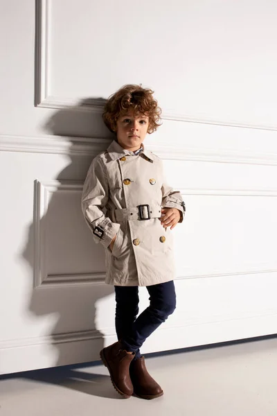 Volledig beeld van een knappe krullende jongen die frontaal poseert in beige jas, kijkend naar de camera. Studiofoto. — Stockfoto