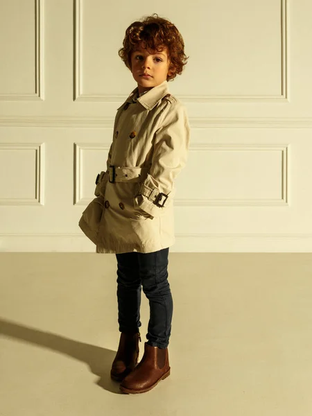 Mooi portret van een krullend jongetje in stijlvolle kleding, met handen in zijn zak, geïsoleerd op een beige achtergrond. — Stockfoto