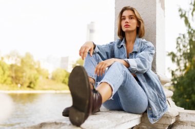 Düşünceli genç bir kızın ön portresi, kot ve bot giymiş, nehrin kenarında oturmuş, başka yöne bakıyor..