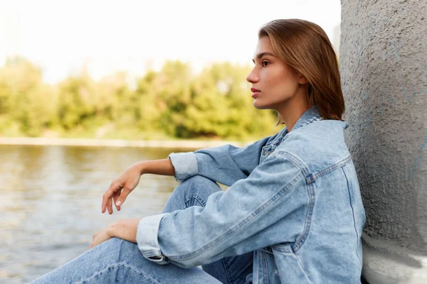 Perfil de uma séria única menina adolescente pensativa, vestida de ganga, sentada fora perto do rio, olhando para longe. — Fotografia de Stock