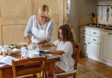 Beyaz önlüklü büyük anne ve güzel torunu mutfakta vakit geçiriyorlar. Büyükanne yeğenine hazırlanmasını öğretir..