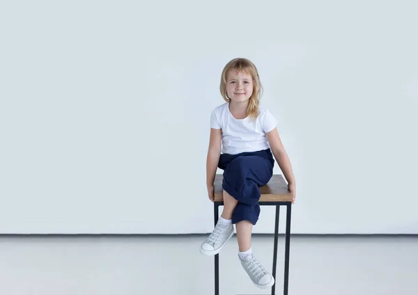 Full längd av söta leende liten flicka med blont kort hår sitter på stol och tittar på kamera, vit bakgrund. — Stockfoto