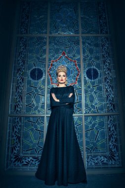 Uzun siyah bir elbise giymiş, uzun boylu sarışın bir kadının resmi. Saç stili, harika bir taç ve küpeleri var..