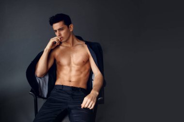 Omzunda ceketi olan, gri arka planda, sandalyede oturan zarif, üstsüz esmer adam..