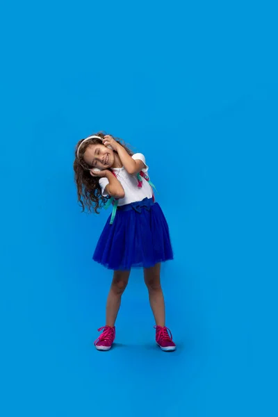 Liten lockig flicka som leker med stora hörlurar, lyssnar på musik och dansar med slutna ögon över blå bakgrund. — Stockfoto