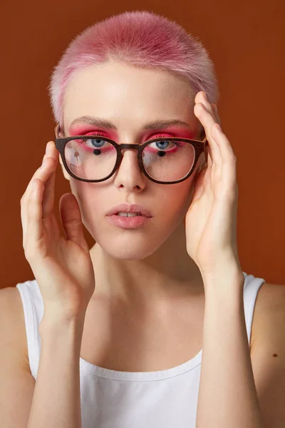 Κοντινό πορτραίτο νεαρής γυναίκας με ροζ μακιγιάζ και κοντές τρίχες με γυαλιά, πάνω από καφέ φόντο. — Φωτογραφία Αρχείου
