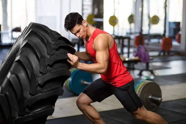 Een sportman in sportkleding duwen immense wiel, geïsoleerd op een wazige sportschool achtergrond. — Stockfoto