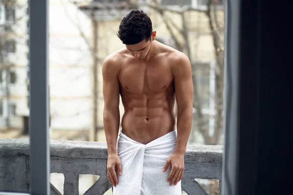 Ein hemdloser muskulöser junger Mann in Badetuch, zeigt Sixpack-Bauch, auf dem Balkon stehend, auf der Straße Hintergrund. — Stockfoto