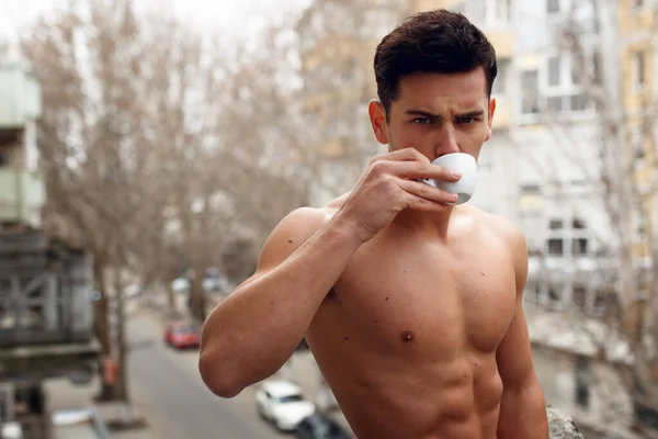 セクシーな、ブルネットの若い男の肖像画バルコニーで彼の朝のコーヒーを飲んで、通りの背景に. — ストック写真