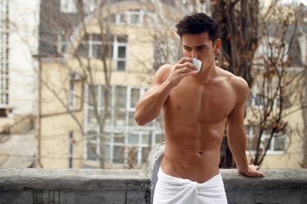 Atractivo morena joven hombre desnudo en toalla de baño, bebiendo una taza de café, posando en el balcón, fondo de la ciudad. —  Fotos de Stock