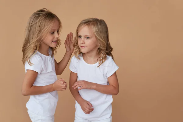 Voorportret van kleine tweelingzusjes met golvend blond haar, met elkaar praten, witte kleren dragen, over beige achtergrond. — Stockfoto