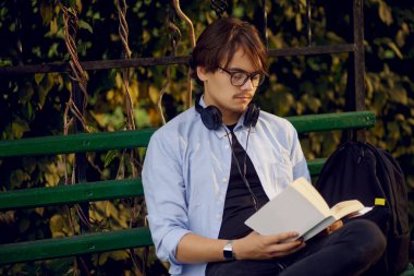 Gözlüklü ve kulaklıklı yakışıklı bir gencin portresi, dışarıda kitap okuyor, şehir parkında izole edilmiş..