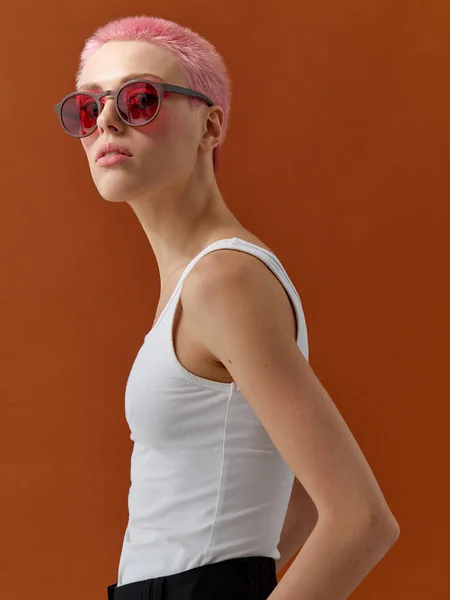 Hipster flicka med korta rosa hår, bär vit t-shirt och svarta jeans och solglasögon med trä fälg och röda linser, över brun bakgrund. Vertikal syn. — Stockfoto